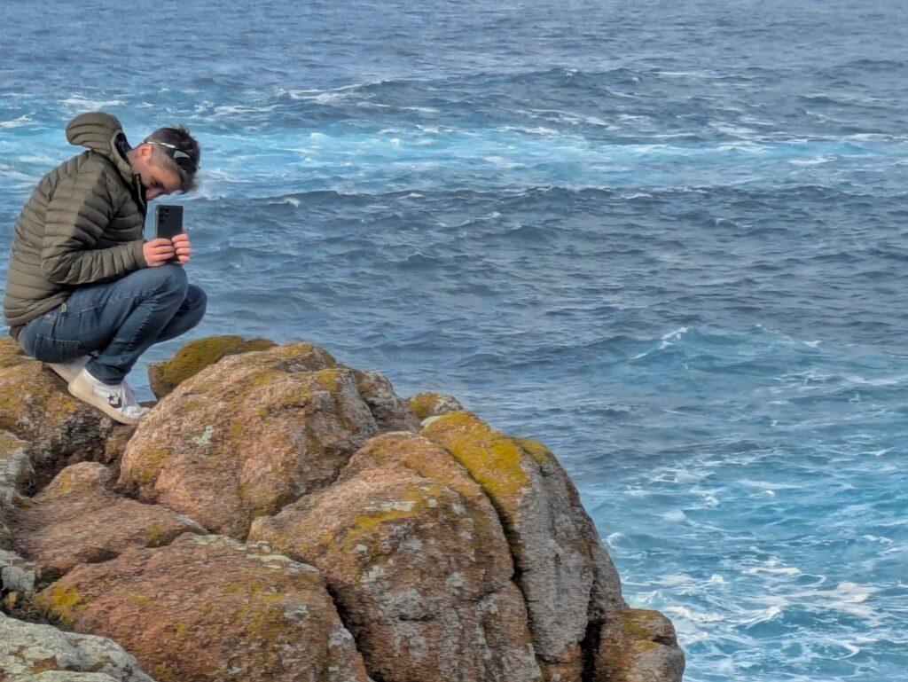 Cass in a green jacket bending down to take a photo on rocks by ocean