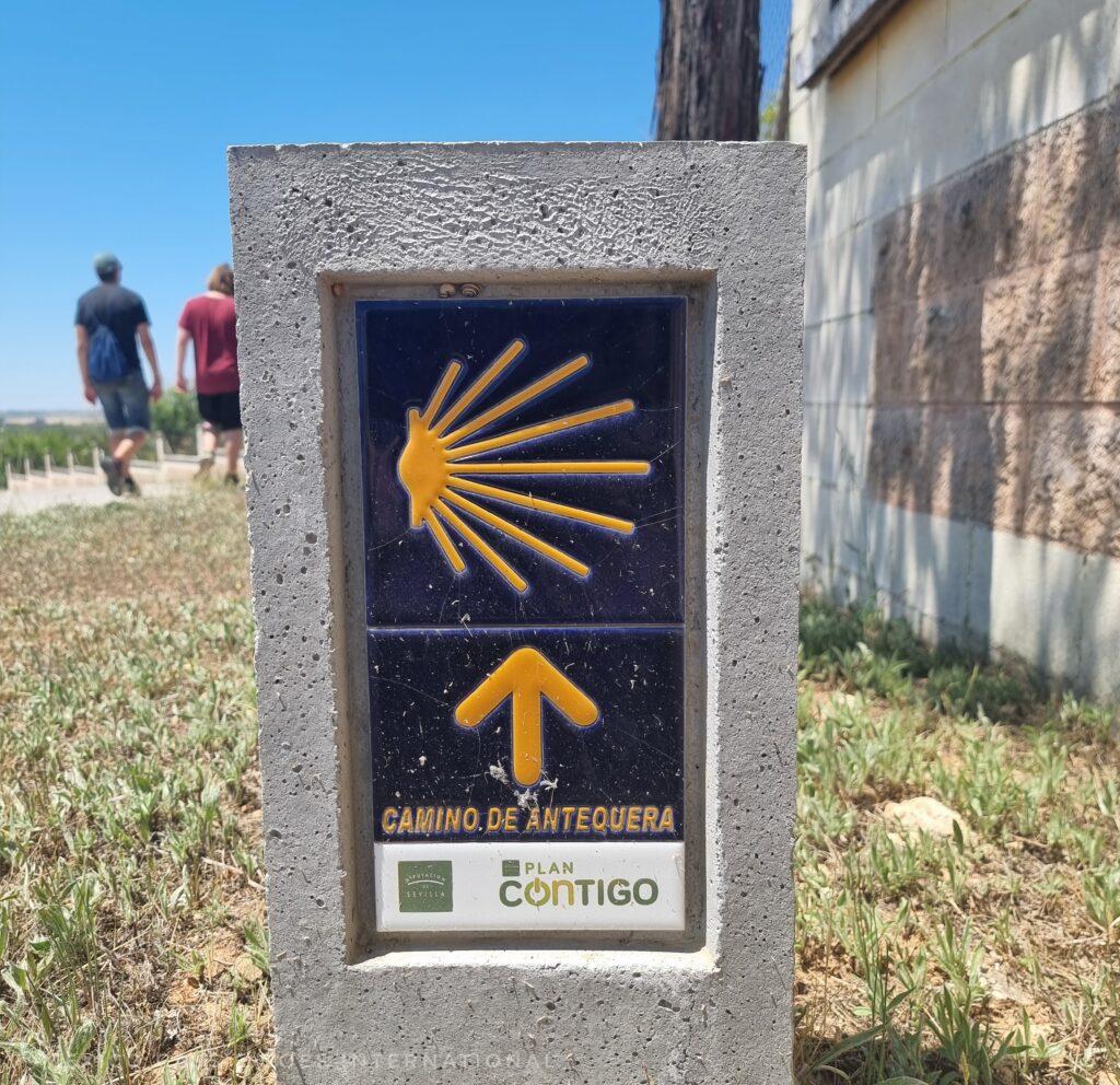 small post with a blue tile with the camino logo in yellow and a yellow arrow, two people in background
