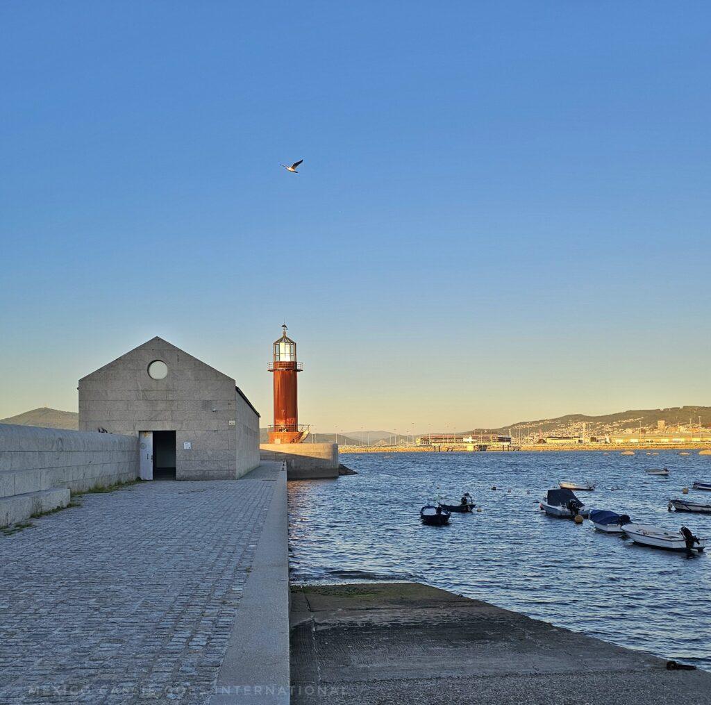 small red lighthouse, white building on left of it and sea with boats on right