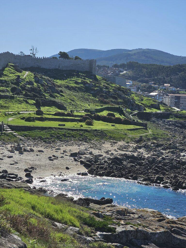 photo of small bay with water that sparkles, beach, green fields, hills and castle