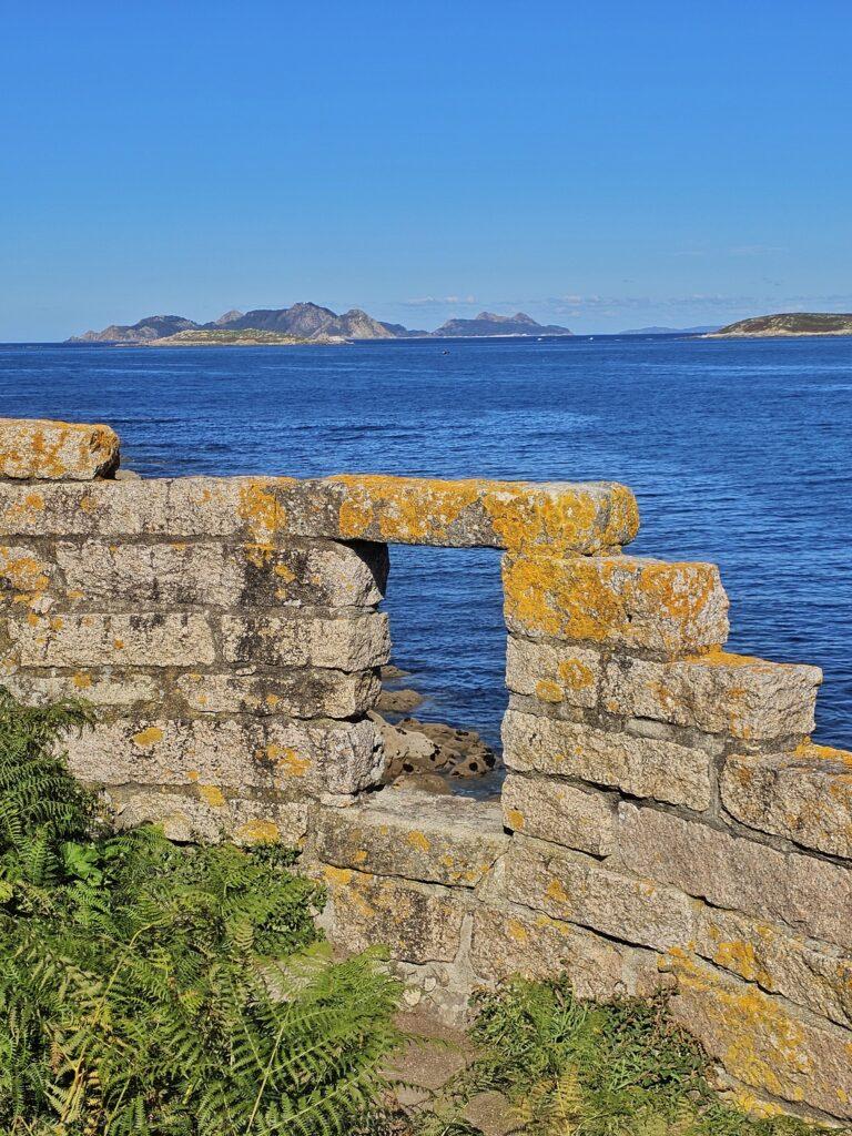 rounded wall, with some yellow moss , wall only goes up a few bricks, window in wall, green plants on left, blue sea and islands behind
