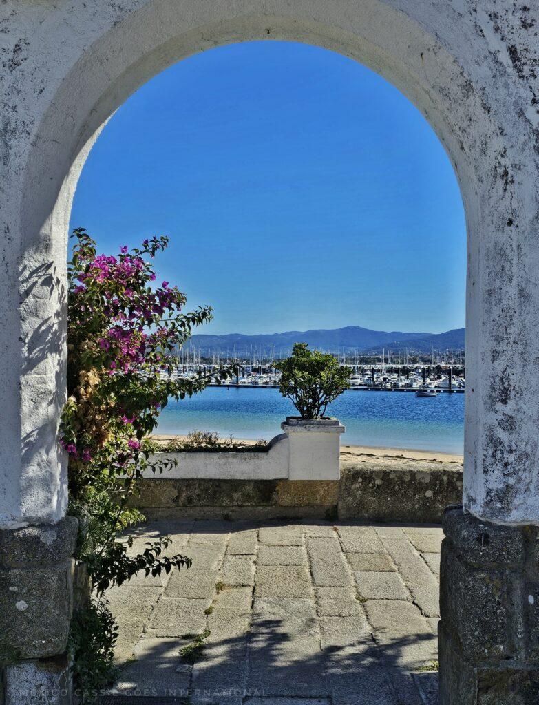 white arch through to path, beach, sea. blue sky and pink plant
